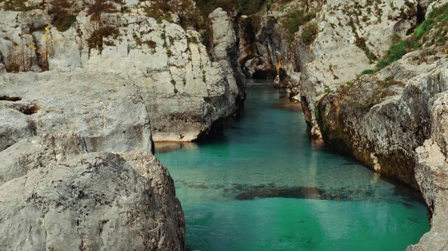 Cinematic shot of the Great Soča Gorge with gentle blue water flowing between canyon walls, rocks, and greenery under bright sunlight in pristine alpine scenery. Lepena Valley.