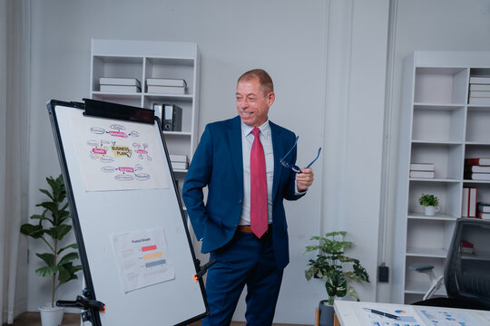 Smiling businessman holding eyeglasses, standing next to a whiteboard with a business plan diagram and product growth charts, presenting concepts in a modern corporate office - Powered by Adobe