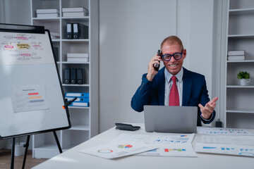 Experienced businessman actively engaging in a phone call, working on a laptop, and analyzing charts and data on his desk in a modern office setup, demonstrating success