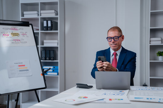 Senior businessman staying connected and handling business tasks, from messaging to reading reports, using a smartphone while sitting at an office desk with a laptop and various documents