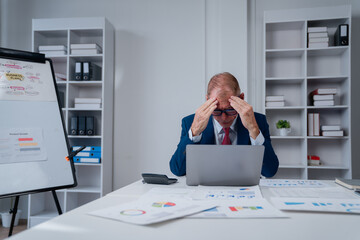 Senior businessman slumped at his office desk with hands on his head, overwhelmed by laptop,...
