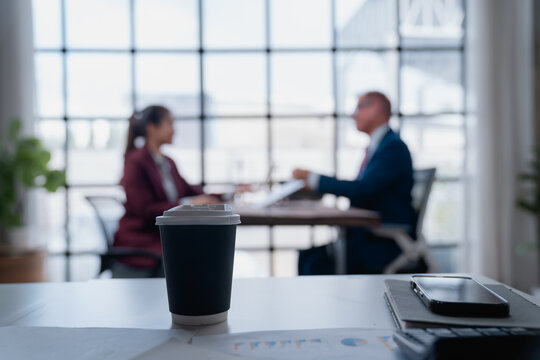 Disposable coffee cup on a meeting room desk, with two blurred business professionals collaborating or discussing work in the background, representing a corporate coffee break or business discussion - Powered by Adobe