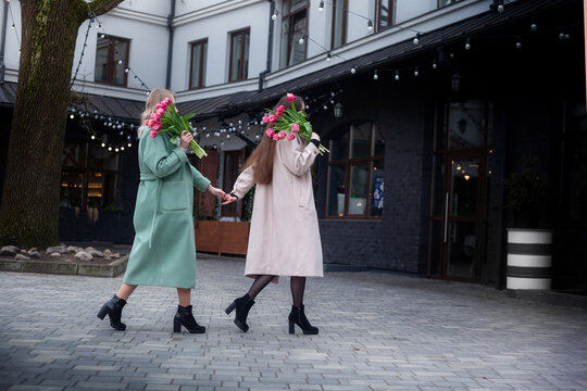 Women in fashionable coats are walking hand in hand, carrying colorful flower bouquets, radiating happiness and friendship in a vibrant outdoor atmosphere