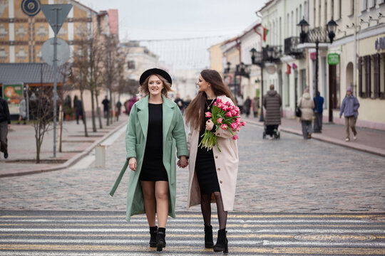 Two young Caucasian women walk together on a city street. One wears a green coat and the other a pink coat, both smiling and enjoying their time. One holds a bouquet of flowers