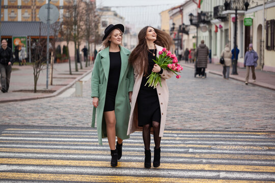 Two young Caucasian women walk together on a city street. One wears a green coat and the other a pink coat, both smiling and enjoying their time. One holds a bouquet of flowers