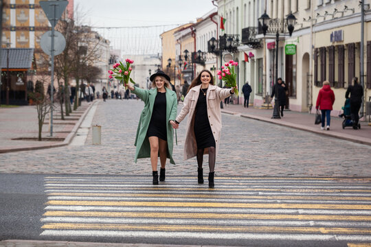 Two young women walk hand in hand across a city crosswalk. They wear stylish coats and hold flowers. The urban street is lively with pedestrians and shops.