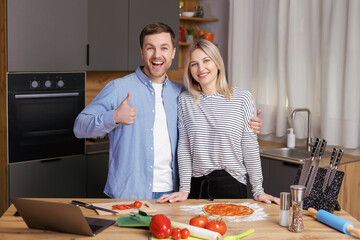 Happy young couple is preparing healthy and tasty food on modern kitchen. Attractive woman and man looking at camera, man gesturing finger up.