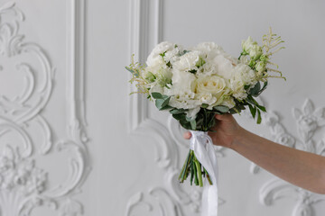 Hand holding a luxurious mixed bridal bouquet with white peonies, cream roses, and assorted greenery against a backdrop of ornate white stucco wall paneling