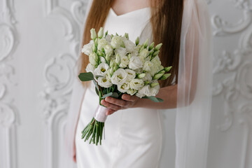 Elegant bride in a white satin gown holding a round bridal bouquet of white eustoma (lisianthus)...