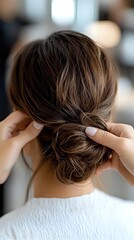 Naklejka premium Professional hairstylist creating elegant low bun hairstyle with brown hair, close-up view from behind showing detailed styling technique and texture.