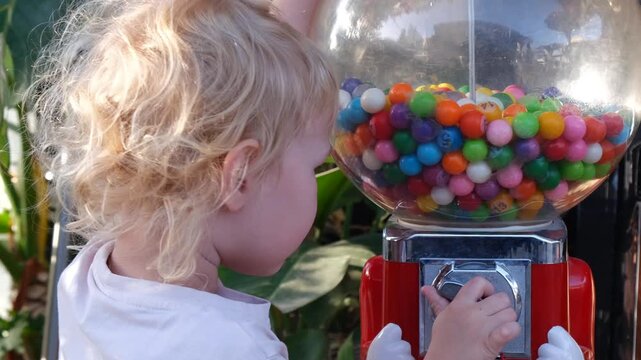 Curious toddler turning the handle of a colorful gumball machine. Bright candy balls in a transparent globe evoke childhood joy and sweet anticipation