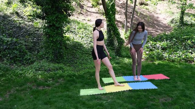 Fitness girlfriends stand on mats in park warming knees gently. Women bend legs rotating joints and chat softly while preparing body for session