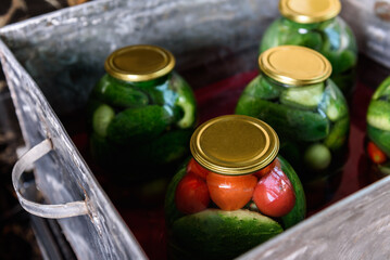 Preservation of season vegetables. A woman sterilizing jars of cucumbers and tomatoes in a large barrel in the kitchen.