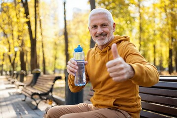 Smiling senior man sitting on park bench holding water bottle and showing thumbs up during autumn sport day.