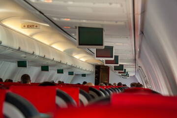 View inside a modern airplane cabin, featuring red seating, overhead storage, and multiple passenger entertainment screens