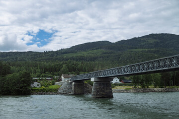 A sturdy metal truss bridge spans a calm river, connecting forested banks under a cloudy sky.