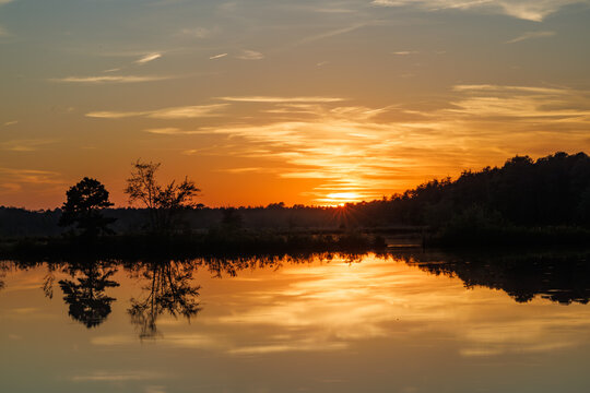 Golden sunset glows above calm Pine Barrens lake, with trees reflecting on still water beneath a warm orange sky at the end of the day.