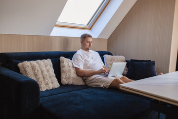 Young man sitting on sofa with laptop and smartphone, working remotely or managing online tasks. Concept of freelancing, digital lifestyle, mobile technology, e-banking, online shopping, communication