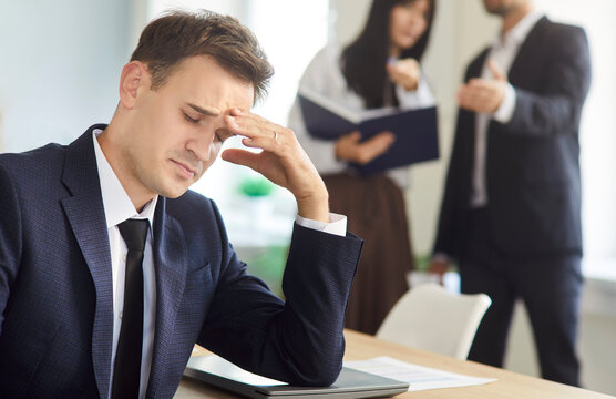 A worried businessman sits at desk under stress and pressure as colleagues argue behind him. Stressed employee faces workplace conflict and criticism. Bullying, harassment, burnout, anxiety.
