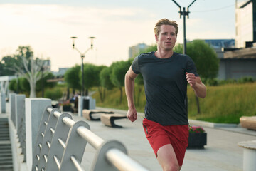 Young man jogging outdoors along urban promenade focused expression athletic build running past metal railing with trees and modern buildings in background