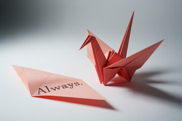 Red origami crane beside folded paper displaying the printed word Always