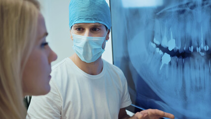 A dentist in surgical attire consults with a patient, pointing at a dental X-ray in a clinical...