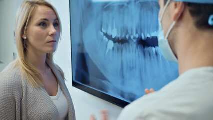 A female patient consults with a dentist, examining a dental X-ray on a monitor in a clinical...