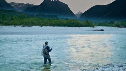 An angler stands in a serene river, fly rod poised, surrounded by lush greenery and majestic mountains, capturing the tranquility of freshwater fishing and the connection with nature.