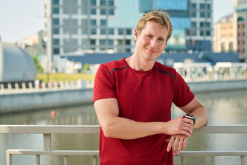 Portrait of Young man smiling and leaning on railing outdoors wearing smartwatch standing in urban area with modern buildings and river in background