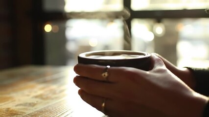 Close-up of a person's hands with rings holding a steaming hot mug of coffee or tea in a cozy cafe. - Powered by Adobe