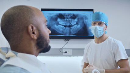A Black patient consults a dentist in a modern clinic. The dentist, wearing a surgical cap and mask, explains an X-ray on a monitor.