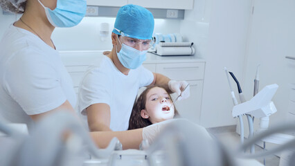 A dentist examines a young patient in a modern, clean dental office. Both wear protective gear, emphasizing hygiene and safety.