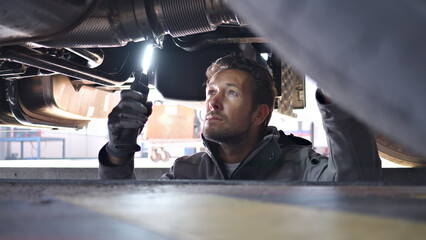 A mechanic inspects a vehicle's undercarriage in a well-lit auto repair shop, using a flashlight to examine engine components with precision and expertise. The professional, clean workshop.