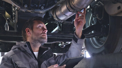 A mechanic examines a car's exhaust system with a tablet in a well-lit auto shop, emphasizing precision and detail.