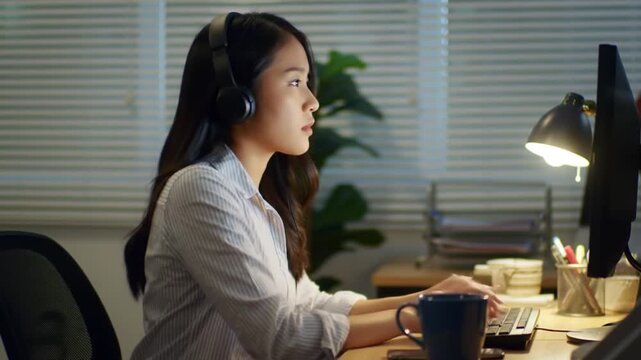 A focused young Asian woman wearing headphones works late at night on her computer in a dimly lit office.