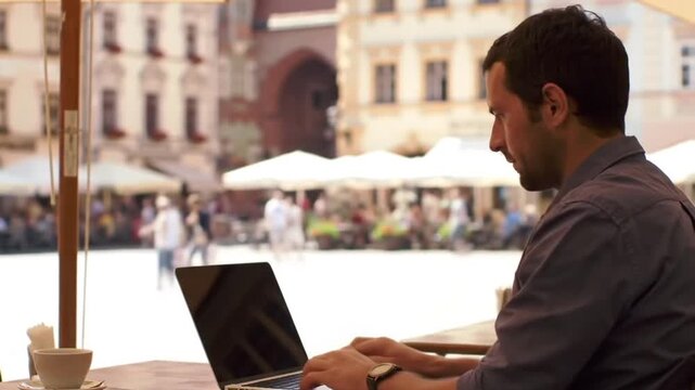 A man works on his laptop at an outdoor cafe in a bustling European city square.