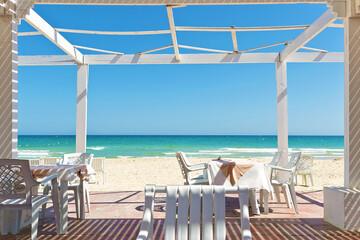 White chairs and tables in wooden pergola on a sandy beach, clear blue sky and calm sea in Tunisia. Gazebo or patio is place for relax on sea shore in restaurant 