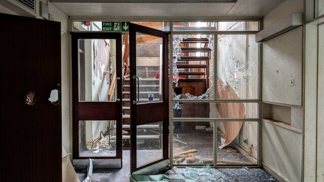 Derelict building stairwell, smashed broken glass door. Doorway floor debris, interior natural light from stairs, shattered windows disaster aftermath. Hurricane, earthquake damaged place staircase.
