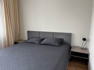 Modern minimalist bedroom interior with a neatly made double bed covered in grey bedding, matching pillows, and simple furniture. Natural light enters through curtains, calm and clean atmosphere.
