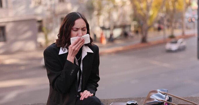 Businesswoman coughs and covers mouth with white tissue in autumn. Woman in business suit sits on sidewalk parapet and coughs into napkin. Slow motion