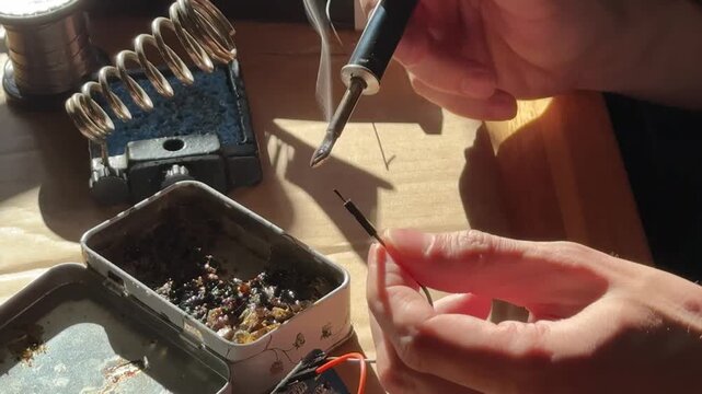 Close-up of skilled hands using a soldering iron to join electrical wires in a workshop. Precision electronics work and technical craftsmanship in an industrial setting.
