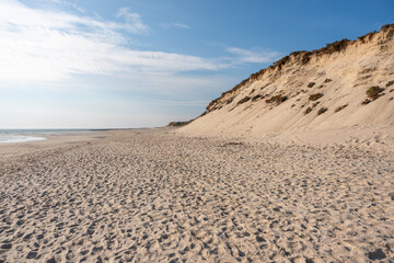 A sandy cliff stretches along the empty beach. The coast meets the calm sea under a bright sky.