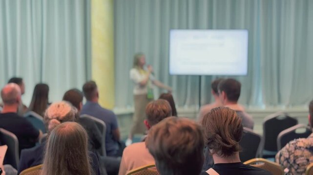 Presenter delivers professional talk to attentive audience at conference setting with large screen projection and seating rows conveying learning, engagement, knowledge sharing
