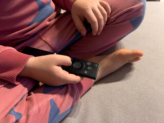 Close-up view of a child holding a TV remote control while sitting on a couch at home. The image illustrates the concept of parental control, screen time limits, and digital monitoring.