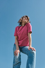 Beautiful carefree young woman with curly hair in pink t-shirt and jeans smiling and looking aside on blue sky background low angle shot.