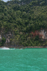 Longtail boat navigating the emerald green waters of Ratchaprapha Dam, surrounded by lush rainforest and towering limestone cliffs in Khao Sok National Park