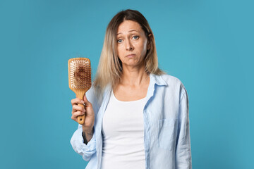 Alopecia problem. Sad woman holding brush with lost hair on light blue background