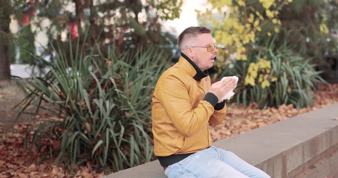 Man in jacket sits on park parapet sneezing into tissue during autumn allergy problem. Man bends forward holding paper napkin near face repeatedly
