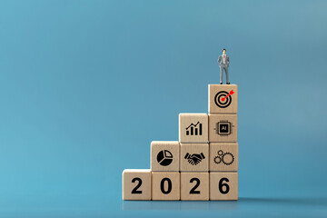 Wooden blocks stacked as a staircase with 2026 typography, business icons chart, handshake, gears, AI, target, and miniature leader on top blue copy space, for strategy, roadmap, KPI and growth goals
