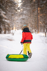 Cute happy kid boy runs through the winter forest and carries a snow inflatable sled. Baby in a red jacket, yellow pants and a blue hat in a snowy winter forest. Activities for children